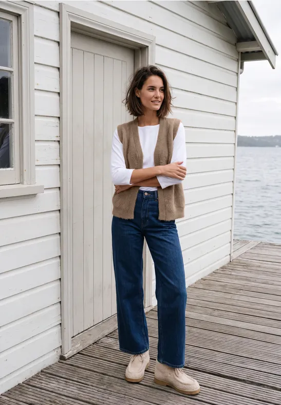 Frau mit Weste und Jeans steht lächelnd auf einem Steg vor einem weißen Haus am Wasser.