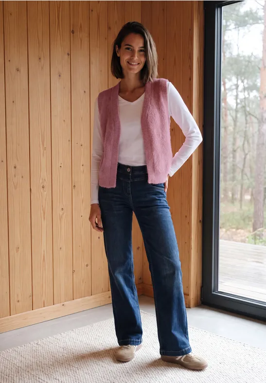 Frau mit rosa Cardigan, weißem Shirt und Jeans steht in Holzraum vor Fenster.