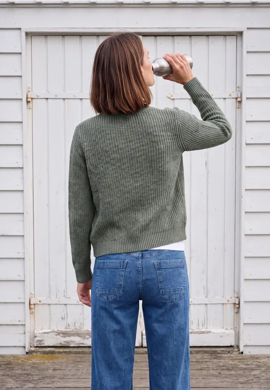 Eine Frau in grünem Pullover und blauer Jeans trinkt aus einer Flasche und steht vor einer weißen Holztür.