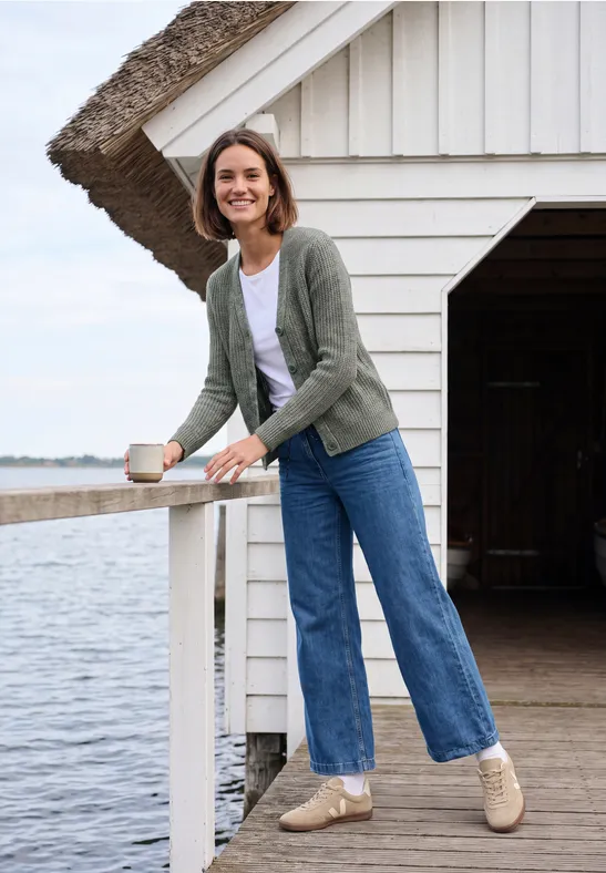 Frau lehnt auf einem Steg an einem Haus am See, hält eine Tasse, lächelt, trägt Jeans und Cardigan.