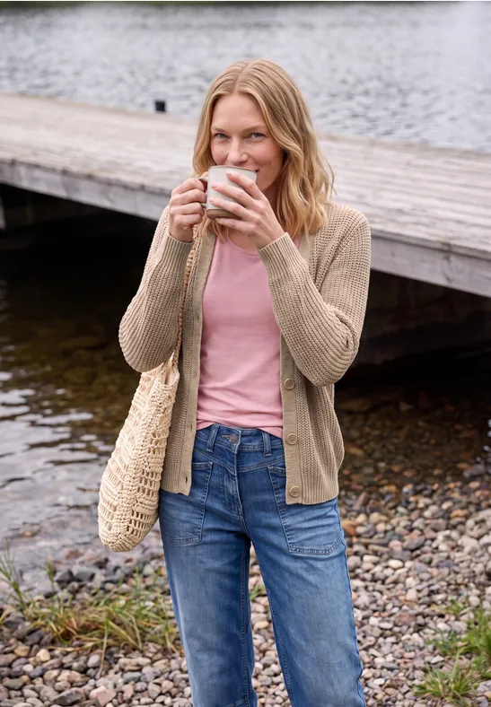 Frau sitzt am Seeufer auf einem Holzsteg, hält eine Tasse, trägt Jeans, rosa Shirt, beige Strickjacke und Korbtasche.