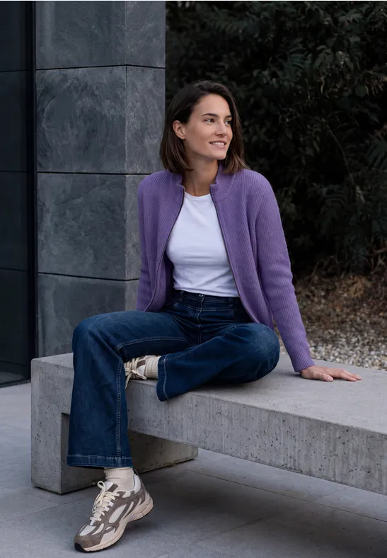 Eine Frau sitzt lächelnd auf einer Betonbank, trägt lila Strickjacke, weiße Bluse, Jeans und Sneaker.