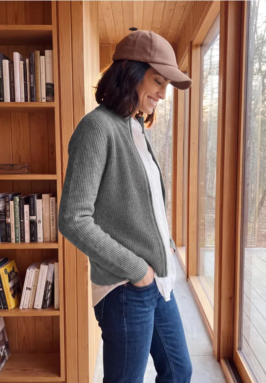 Frau in grauem Cardigan und Baseballcap steht entspannt am Fenster vor Bücherregal und blickt nach draußen.