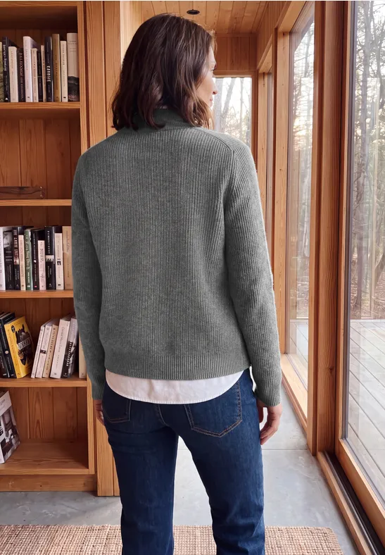 Person im grauen Pullover und Jeans steht vor Holzregal und großen Fenstern und blickt hinaus.