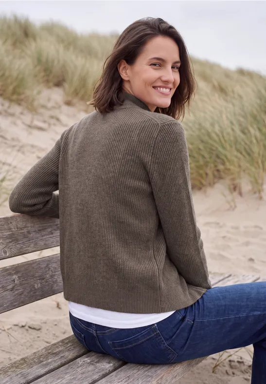Frau sitzt lächelnd auf einer Bank am Strand, trägt grüne Jacke und Jeans vor Dünen mit Strandgras.