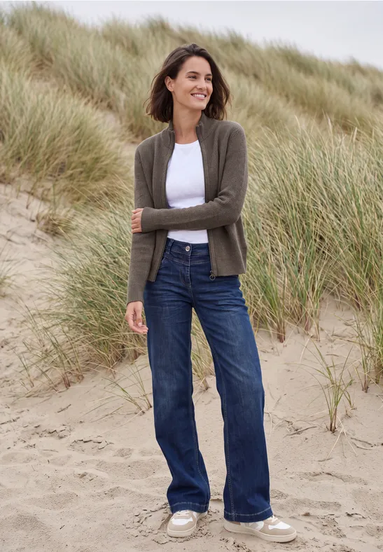 Frau im weißen Shirt, grüner Strickjacke und blauen Jeans steht lächelnd in Dünengras am Strand.