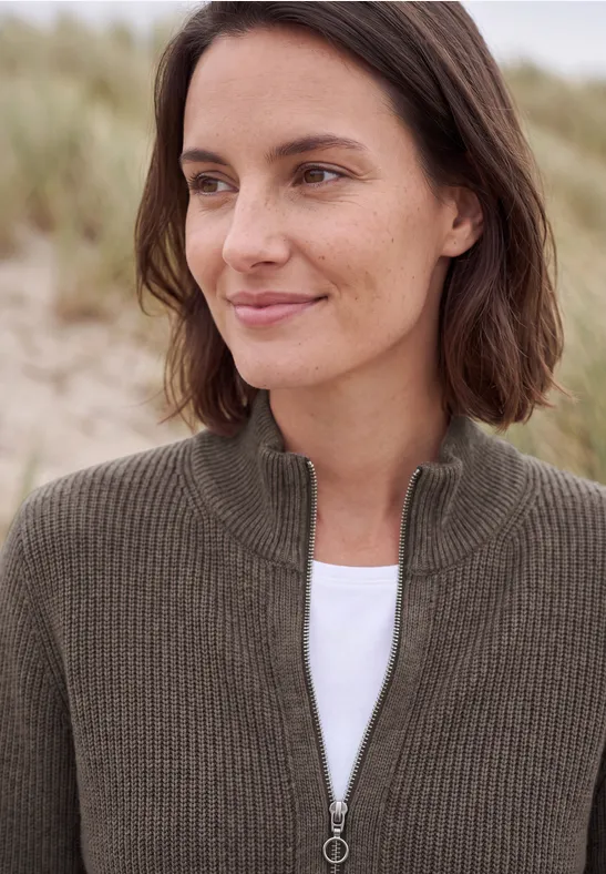 Eine Frau mit braunem Strickpullover und weißem T-Shirt vor Sanddünen am Strand.
