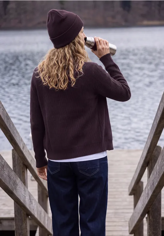 Rückansicht einer Person mit Mütze, Pullover und Jeans, die auf einem Steg am See aus einer Flasche trinkt.