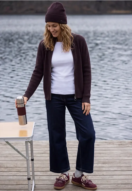 Frau steht auf Holzsteg am See, trägt Mütze, weißes Shirt, Strickjacke und weite Jeans, gießt Getränk aus Thermoskanne.