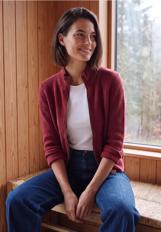 Vrouw in een rood vest, wit shirt en jeans zit op de vensterbank, kijkend naar buiten.