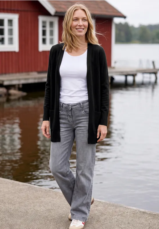 Eine Frau in Jeans und Cardigan steht auf einem Steg vor einem roten Holzhaus am See.