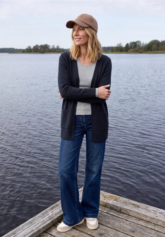 Eine lächelnde Frau in Baseballcap, Cardi, Jeans und Sneakers steht auf einem Steg vor ruhigem See.
