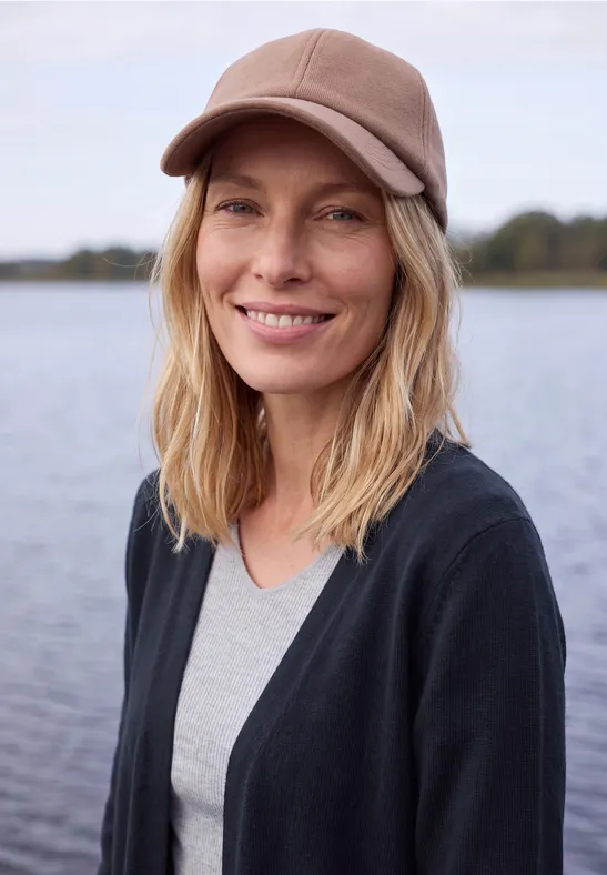 Eine blonde Frau mit Kappe lächelt entspannt vor einem See.
