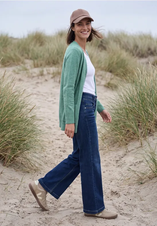 Frau in grünem Cardigan, Jeans und Cap steht lächelnd in Dünen mit Strandgras.