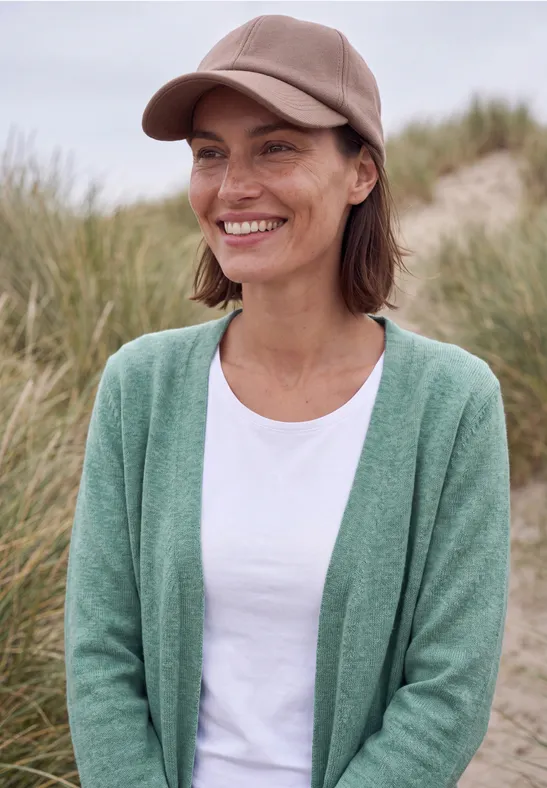 Eine lächelnde Frau trägt eine grüne Strickjacke, weißes T-Shirt und eine beige Mütze an einem Dünenstrand.