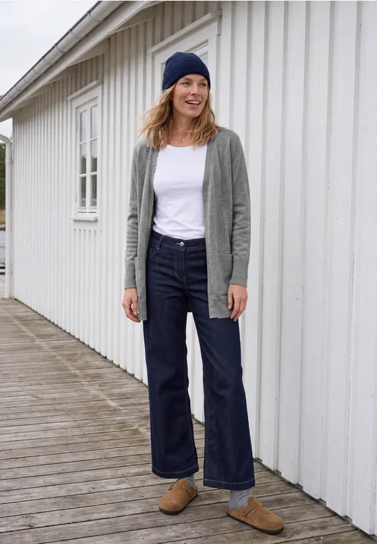 Frau in lässigem Outfit (Beanie, Cardigan, weißes T-Shirt, weite Jeans, Loafers) vor weißem Holzhaus.