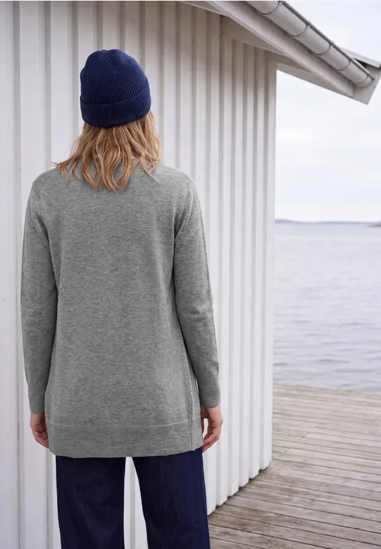 Person mit grauem Pullover und blauer Mütze steht rückwärts auf Holzsteg am Wasser.