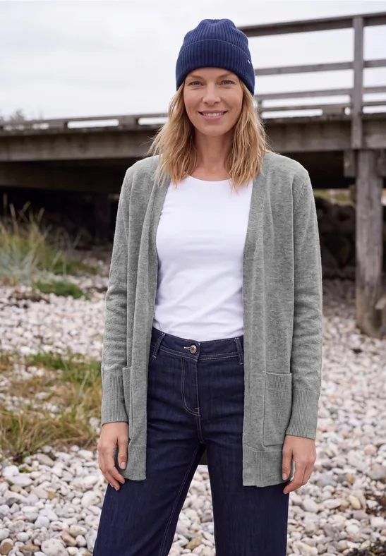Eine lächelnde Frau im grauen Cardigan, weißem Shirt, Jeans und blauer Mütze steht entspannt am steinigen Ufer vor einem Holzsteg.