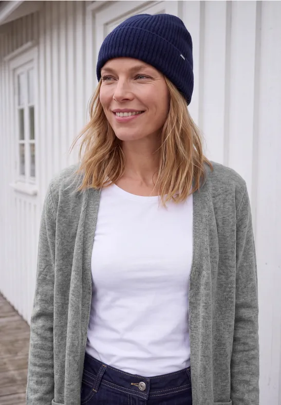 Eine lächelnde Frau in Strickmütze, weißem Shirt und grauem Cardigan steht vor hellem Holzhaus.