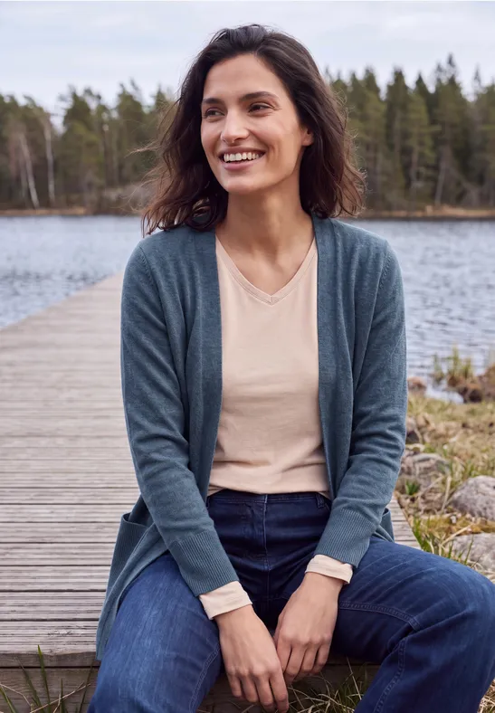 Frau sitzt lächelnd auf Holzsteg am See, trägt Cardigan und Jeans vor bewaldetem Hintergrund.