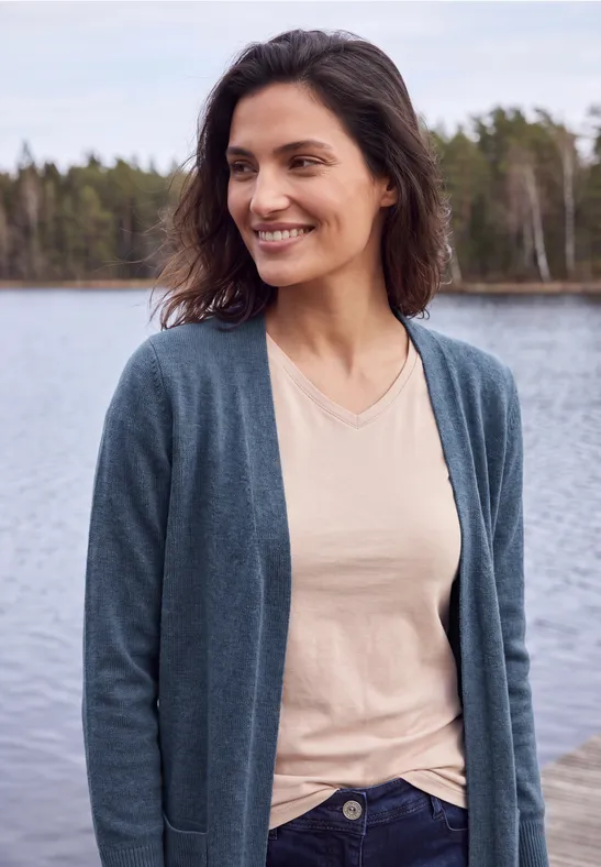 Eine lächelnde Frau am Seeufer trägt eine Strickjacke über einem hellen Shirt.