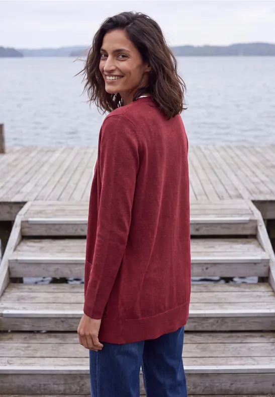 Eine Frau in rotem Cardigan und Jeans steht lächelnd auf einem Holzsteg am Wasser.