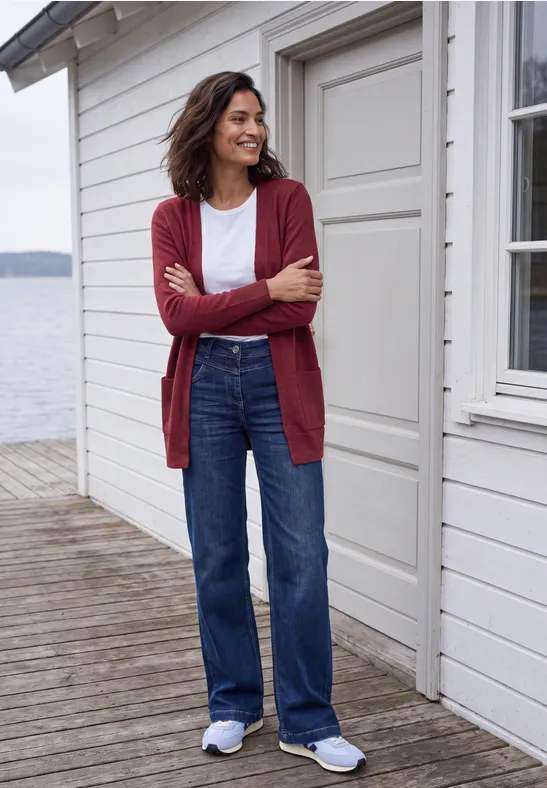 Frau in rotem Cardigan, weißem Shirt und blauen Jeans lehnt lächelnd an Holzwand auf einer Seeterrasse.