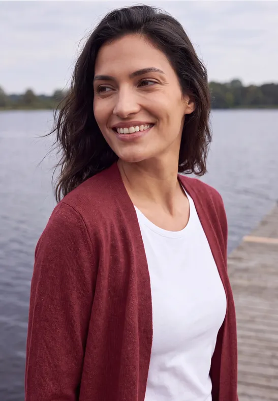 Eine lächelnde Frau im weißen T-Shirt und roter Strickjacke steht an einem Steg am See.