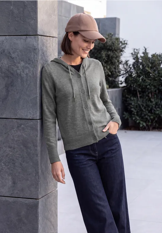 Une jeune femme en sweat à capuche vert, jean et casquette, appuyée et souriante contre un mur en béton.
