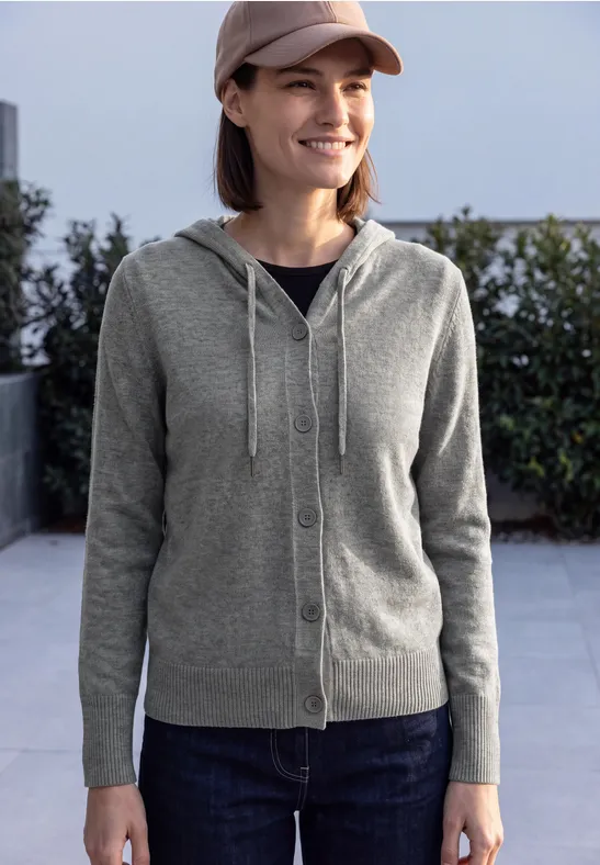 Femme souriante en cardigan gris à capuche, jean et casquette, debout dehors devant une haie.