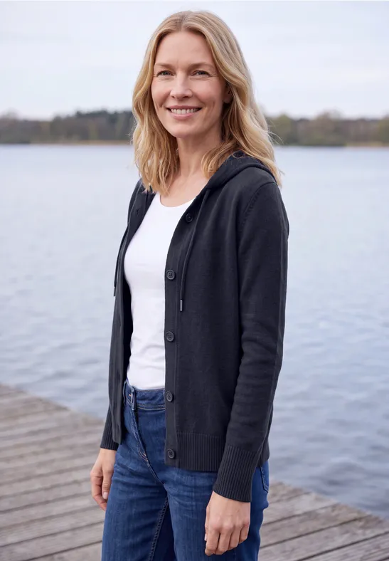 Eine Frau steht lächelnd an einem Steg am See, trägt weiße Bluse, dunkle Jacke und blaue Jeans.