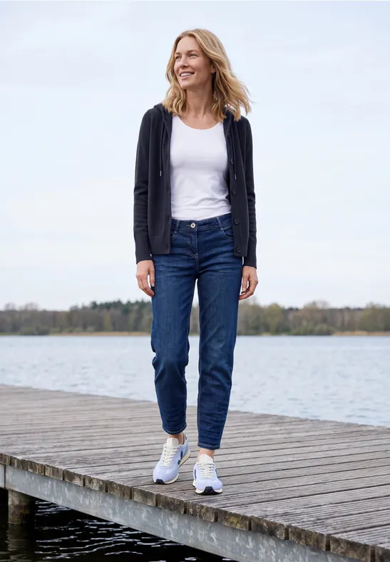 Frau läuft auf einem Steg am See, trägt Jeans, weißes Top und Cardigan