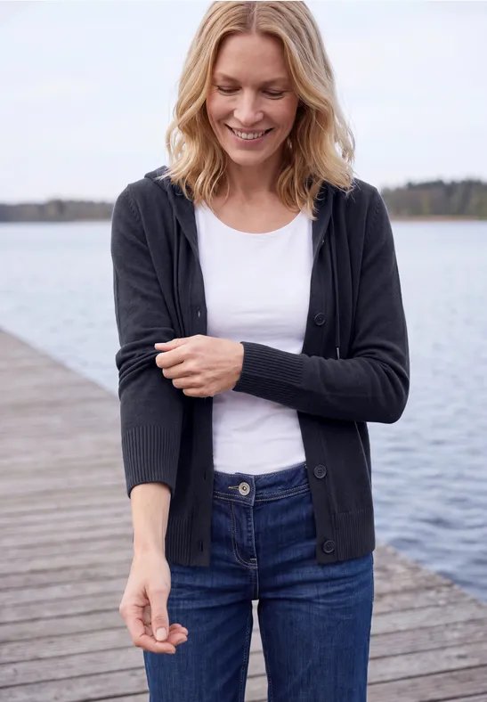 Frau steht lächelnd auf einem Holzsteg am See, trägt Jeans, weißes Shirt und schwarze Strickjacke.
