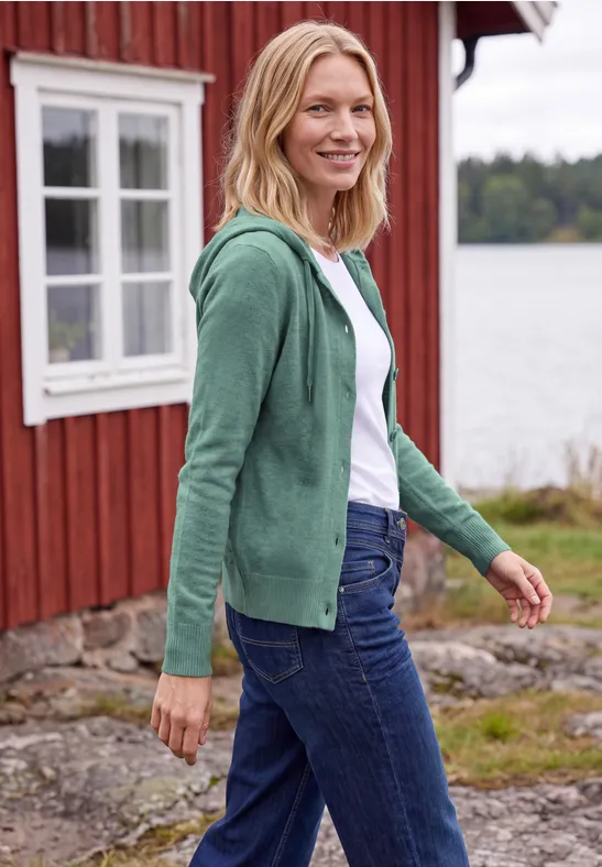 Een blonde vrouw in een groene capuchontrui en jeans lacht voor een rood houten huis aan een meer.