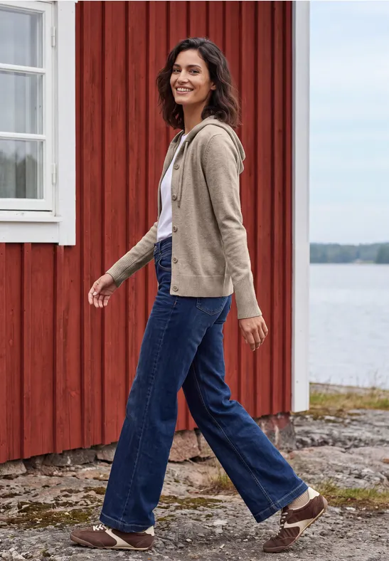 Frau in Jeans und Jacke spaziert fröhlich an einem roten Holzhaus am Wasser.