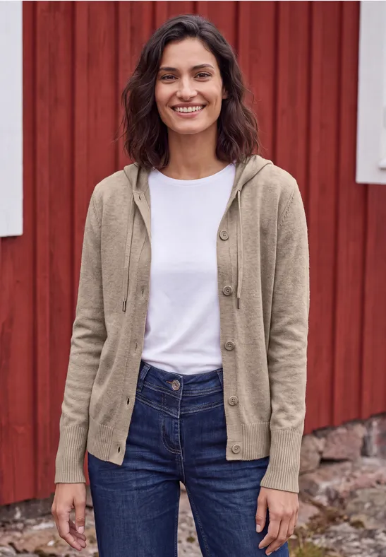 Eine lächelnde Frau in Jeans, weißem T-Shirt und beiger Strickjacke vor einer roten Holzwand.