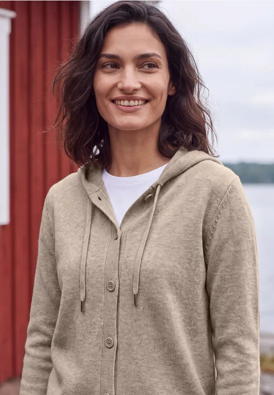 Eine lächelnde Frau in beige Kapuzenjacke und weißem Shirt steht draußen vor einem roten Holzhaus am Wasser.