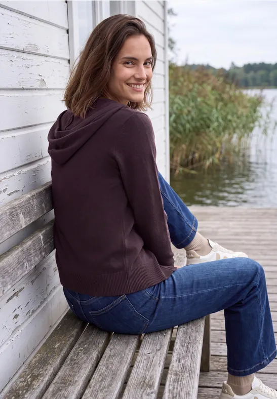 Frau mit brauner Kapuzenjacke und Jeans sitzt seitlich auf einer Holzbank an einem See und lächelt.