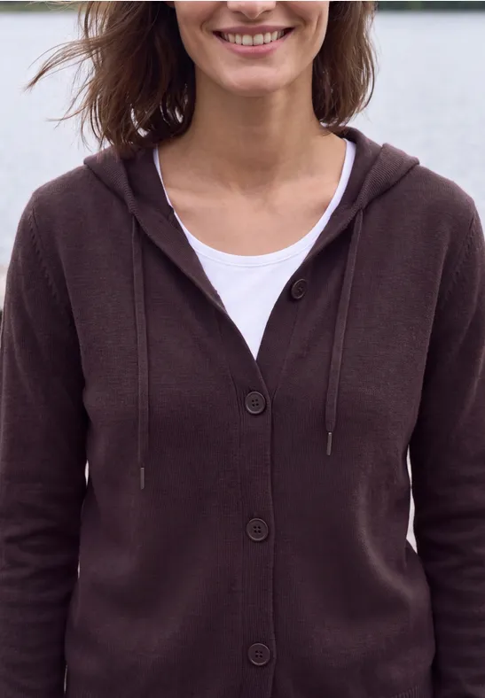 Lächelnde Frau in braunem Kapuzenpullover und weißem Shirt vor unscharfem Hintergrund.