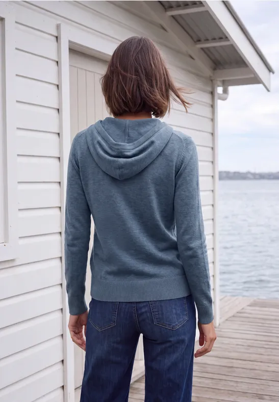 Person in Jeans und Kapuzenpullover steht auf einem Steg neben einem Bootshaus und blickt auf das Wasser.