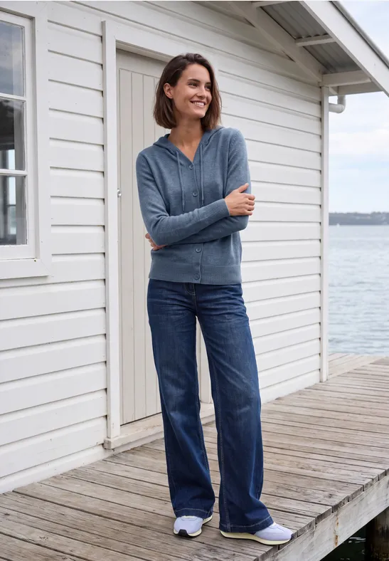 Eine lächelnde Frau in Jeans und Strickjacke steht auf einem Holzsteg vor einem weißen Haus am Wasser.