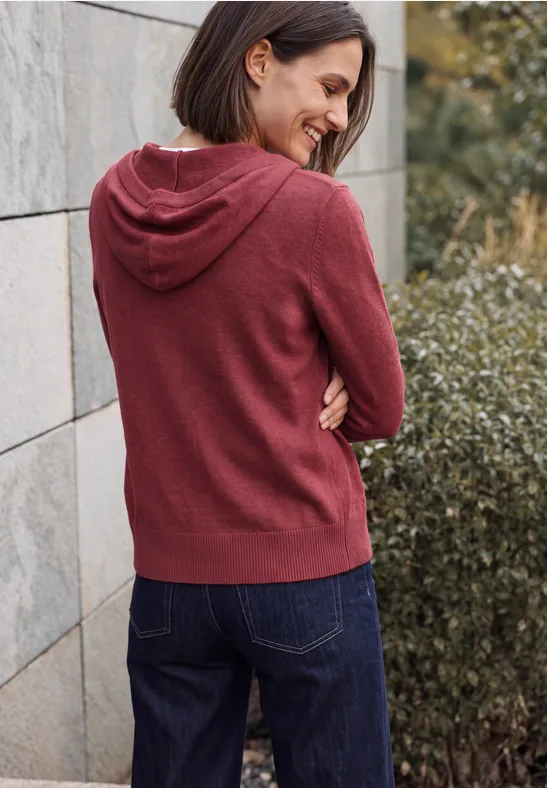 Femme souriante vue de dos, portant un sweat à capuche bordeaux et un jean, appuyée sur un mur près d’un buisson.