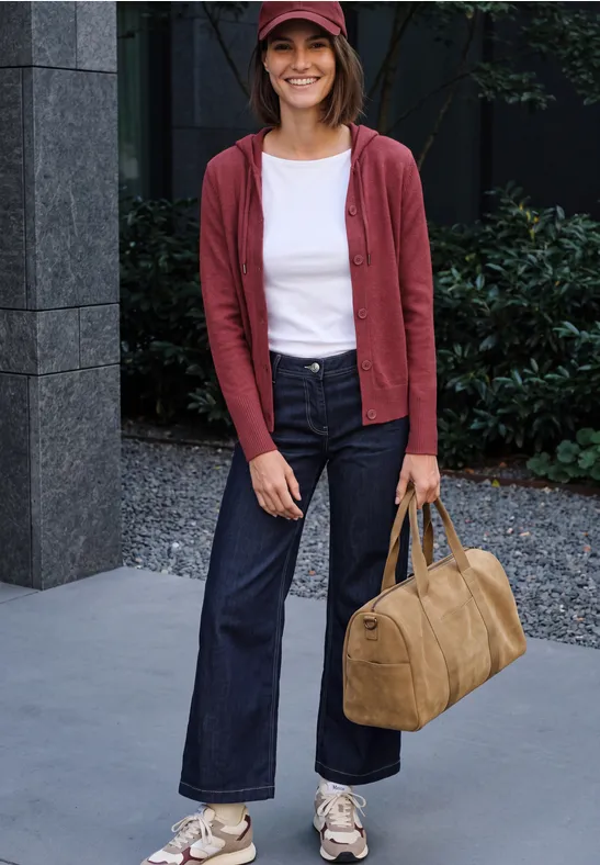 Jeune femme en veste rouge, jean et casquette, souriante, portant un sac beige devant un bâtiment.