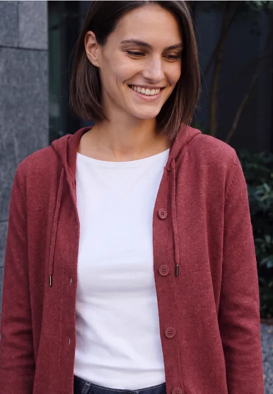 Femme aux cheveux bruns courts, sweat à capuche rouge et t-shirt blanc, souriante et détendue.