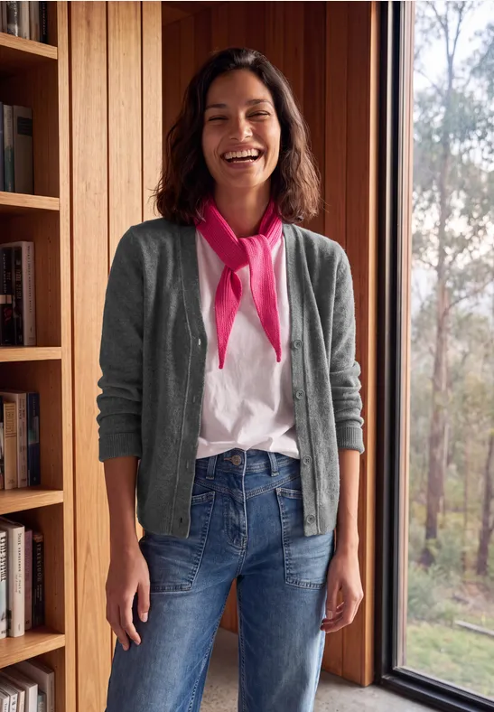 Eine lächelnde Frau in Jeans, T-Shirt und grauer Strickjacke steht locker vor einem Fenster im Holzinterieur.