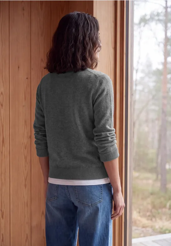 Eine Person mit grauem Pullover und Jeans steht im Holzraum und blickt aus dem Fenster in den Wald.