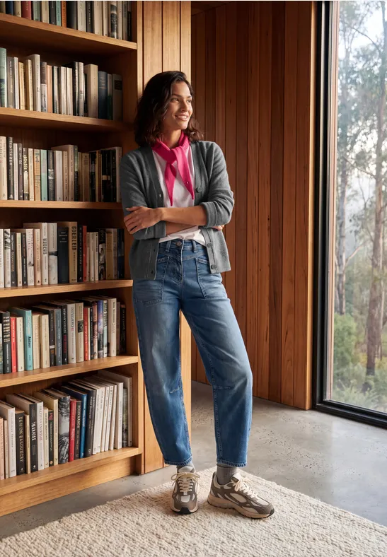 Eine Frau steht lässig vor einem Bücherregal aus Holz, trägt Jeans, Cardigan und blickt aus dem Fenster.
