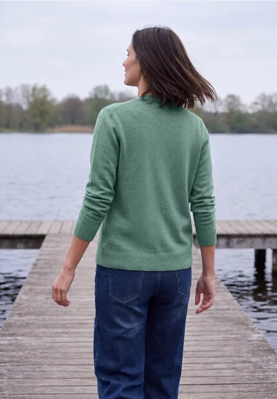 Frau in grünem Pullover und Jeans steht auf Steg und blickt auf ruhigen See.