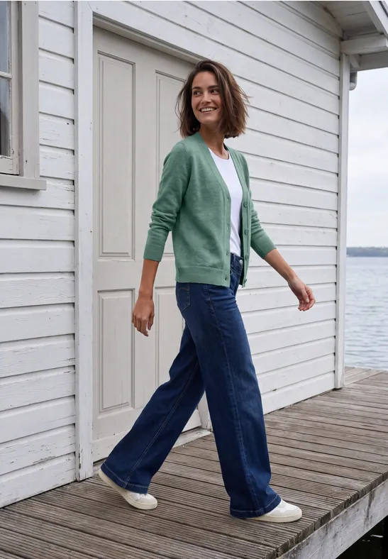 Frau in grünem Cardigan und Jeans spaziert auf einem Holzsteg vor weißem Haus am Wasser.
