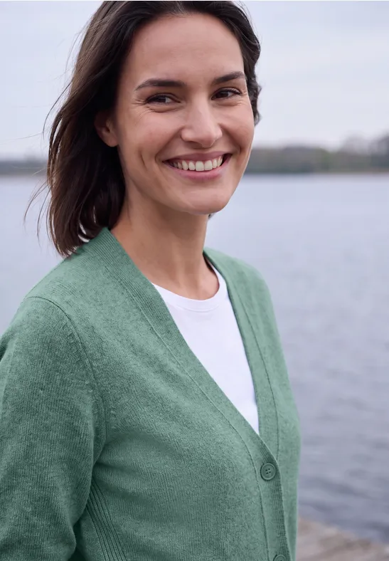 Lächelnde Frau in grünem Pullover vor einer ruhigen Wasserlandschaft.